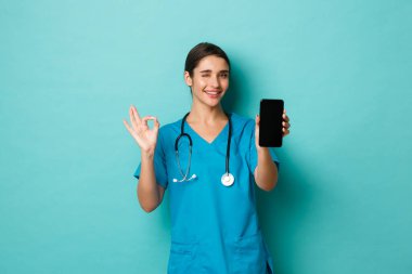 Covid-19, salgın hastalık ve ilaç konsepti. Gülümseyen güzel bir kadın doktor resmi, önlük giyiyor, cep telefonu ekranını gösteriyor ve tamam işareti, tavsiye uygulaması, mavi arka plan