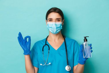 Covid-19 ve karantina kavramı. Tıbbi maskeli, eldivenli ve önlüklü kendine güvenen kadın doktorlara yakın plan. Koronavirüsten korunmak için el dezenfektanı kullanın.