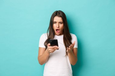 Beyaz tişörtlü, şok olmuş modern bir kızın portresi, akıllı telefon ekranına dehşete düşmüş, mavi arka planda duruyor.
