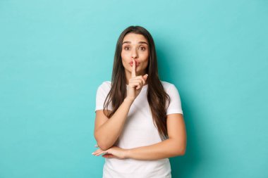 Beyaz tişörtlü tatlı esmer kızın portresi, parmaklarını dudaklarına bastırmış, bir sır söylüyor, sessiz kalmak istiyor, mavi arka planda duruyor.