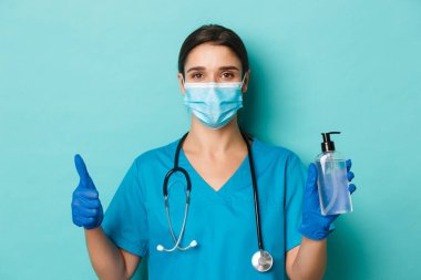Covid-19 ve karantina kavramı. Tıbbi maskeli, eldivenli ve önlüklü güzel kadın doktorlara yakın çekim. Baş parmaklarını gösteren önlükler. Coronavirüs 'ten korunmak için el dezenfektanı kullanma tavsiyesi.
