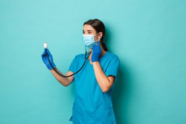 Covid-19, salgın hastalık ve ilaç konsepti. Tıbbi maskeli, eldivenli ve önlüklü güzel bir kadın doktor. Hastanın steteskopla kontrol etmesi, akciğerleri kontrol etmesi, mavi arka planda durması.