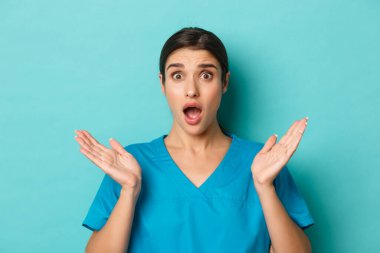 Coronavirus, sosyal uzaklık ve sağlık konsepti. Telaşlı ve endişeli kadın doktorların yakın plan, ellerini yana aç ve çenelerini indir, kameraya endişeli bir şekilde bak, mavi arka planda dur.
