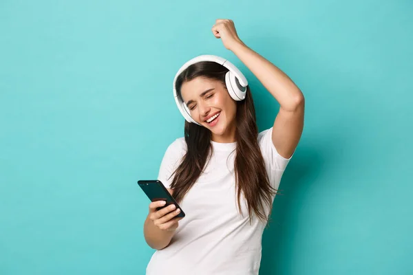 Retrato de la chica bonita moderna en camiseta blanca, escuchando música en auriculares inalámbricos, sosteniendo el teléfono móvil y bailando, de pie sobre fondo azul — Foto de Stock