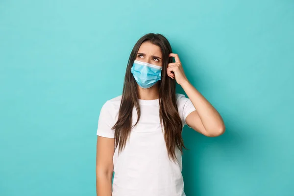 Begreppet pandemi, covid-19 och socialt avstånd. Bild av förbryllad ung kvinna i vit t-shirt och medicinsk mask, ser förvirrad på övre vänstra hörnet, klia huvudet, blå bakgrund — Stockfoto