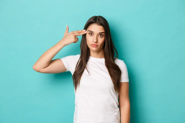 Imagen de modelo femenina molesta y molesta, con camiseta blanca, haciendo letrero de arma de mano cerca de la cabeza, disparándose a sí misma de la angustia, de pie sobre fondo azul —  Fotos de Stock