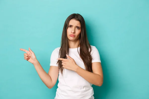 Portrait of sad and disappointed brunette girl in white t-shirt, frowning upset, pointing fingers left at copy space, standing over blue background — Stock Photo, Image