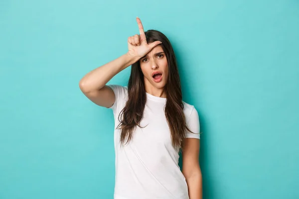 Image of arrogant and sassy brunette woman looking cool, mocking someone, showing loser sign on forehead, standing over blue background — Stock Photo, Image