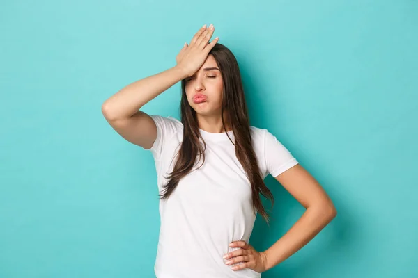 Giovane donna stanca in t-shirt bianca, sospirando e facendo facepalm per esprimere fastidio, sentirsi stufo e teso, in piedi su sfondo blu — Foto Stock