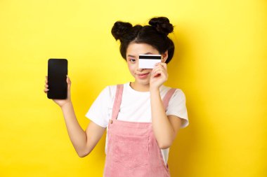 İnternetten alışveriş konsepti. Plastik kredi kartıyla ödeme yapan sevimli Asyalı kız, boş bir akıllı telefon ekranı ve gülümsüyor, sarı arka plan.