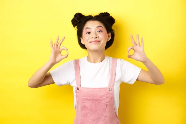 Feliz chica coreana adolescente con maquillaje de belleza, mostrando signos de bien y sonriendo satisfecho, recomendando producto, alabanza buena opción, de pie sobre fondo amarillo —  Fotos de Stock