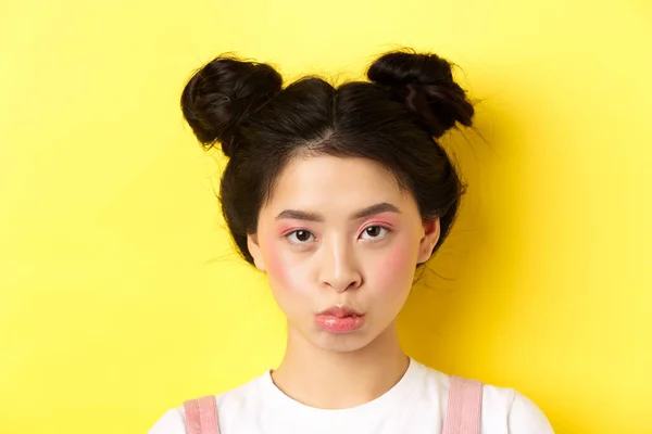 Крупный план портрет угрюмой азиатской девушки дуться и смотреть расстроен на камеру, стоя с гламурным макияжем и парикмахерские на желтом фоне — стоковое фото