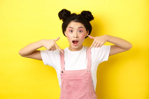 Emocionado adolescente asiático chica con belleza maquillaje, señalando los dedos hacia abajo y la boca abierta fascinado, de pie feliz sobre fondo amarillo —  Fotos de Stock