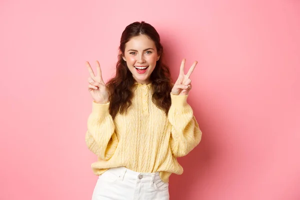 Retrato de modelo feminino morena atraente, sorrindo e mostrando sinais de paz, fazendo gesto v-sign, de pé positivo e feliz contra fundo rosa — Fotografia de Stock