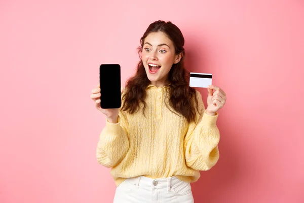 Технологии и интернет-магазины. Захватывающая привлекательная девушка с экраном смартфона, пластиковой кредиткой, с удивлением смотрит на телефон, стоя на розовом фоне — стоковое фото