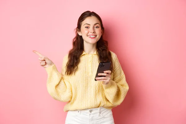 Vacker modern flicka som håller smartphone och peka finger åt sidan, visar PR-text på copyspace när du använder mobiltelefon, står över rosa bakgrund — Stockfoto