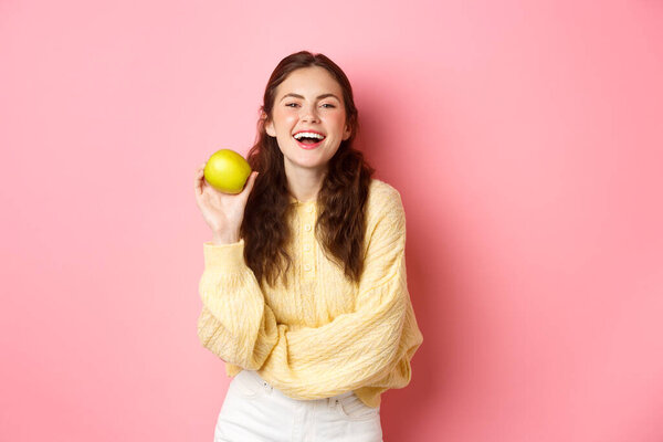 Healthy diet, people and lifestyle concept. An apple day keeps doctor away, girl holding delicious fruit and smiling happy at camera, standing against pink background