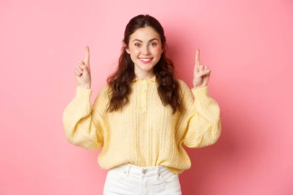Εικόνα ελκυστικής νεαρής καυκάσιας γυναίκας που δείχνει τα δάχτυλα ψηλά, δείχνει διαφήμιση, χαμογελά στην κάμερα, στέκεται πάνω σε ροζ φόντο — Φωτογραφία Αρχείου