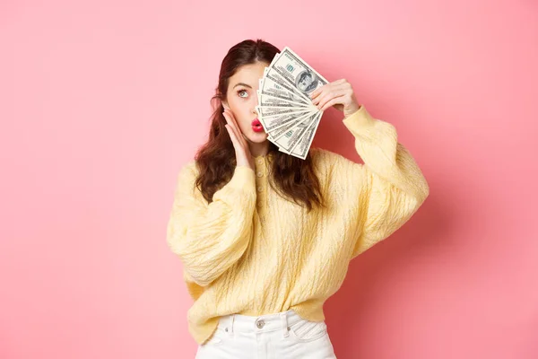 Atractiva modelo femenina mostrando dinero, sosteniendo billetes de dólar en la mitad de la cara, tocando la mejilla y haciendo labios besándose, de pie sobre fondo rosa — Foto de Stock