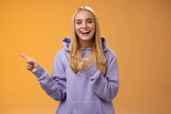 Charismatic firendly good-looking blond young female student pointing left discussing awesome new restaurant suggesting friend go check out laughing smiling happily, orange background