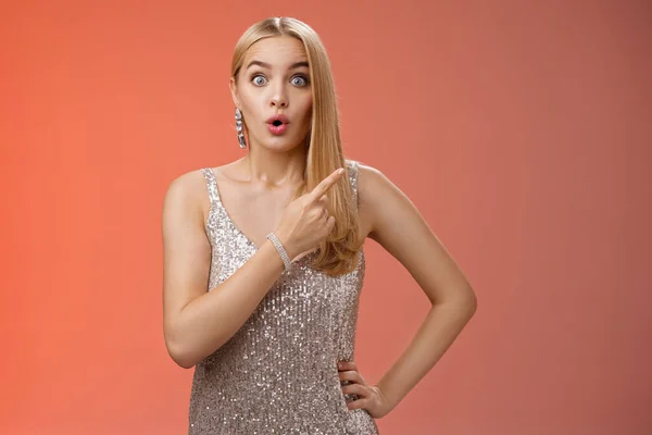 Wondered amused attractive blond woman talking party widen eyes say wow pointing questioned right direction see famous person standing surprised impressed gaze camera, red background
