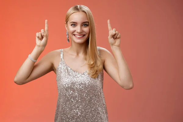 Opgewonden prachtige blonde Europese vrouw in zilver glanzende elegante jurk verhogen handen wijzend omhoog met indrukwekkende ongelooflijke reclame glimlachen gelukkig blij enthousiast willen nemen van dichterbij te kijken, rode achtergrond — Stockfoto