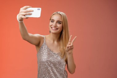 Çekici, genç, narin, sarışın 25 'li yaşlarda, gümüş renkli şık elbiseli, selfie çeken, kolunu uzatan, barış jesti akıllı telefon görüntüsü sunan, internet hayranlarını selamlayan, kırmızı arka planda dikilen genç bir kız.