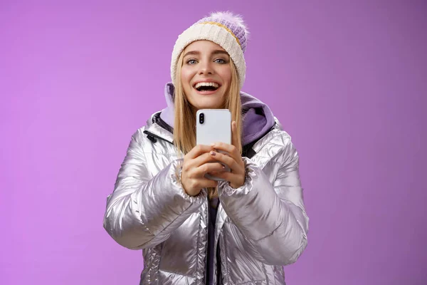 Amüsiert aufgeregt attraktive blonde Freundin hält Smartphone hoch Aufnahme Video Freund Schritt Snowboard zum ersten Mal Erfassung von Erinnerungen mobile Kamera steht glücklich lila Hintergrund — Stockfoto