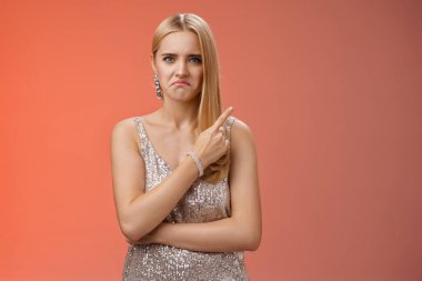 Jealous displeased angry young revengeful blond ex-girlfriend in luxurious silver shiny dress frowning pouting pointing upper right corner displeased pissed standing fed up upset red background