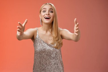 Heyecanlı cazibeli genç sarışın bayana dokundu gümüş parti cazibesi elbisesiyle kameraya doğru ellerini uzattı sarılmak istedi sevimli yavru köpek kollarını tuttu, kırmızı arka planda durdu.