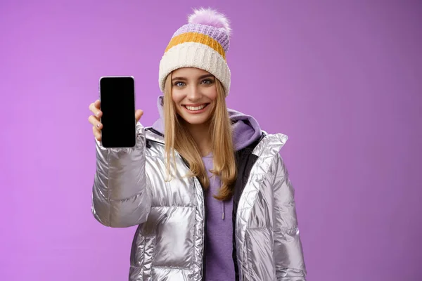 Freundlich fröhlich selbstbewusst blondes Mädchen in silberner stylischer Winterjacke Hut ausstrecken Arm zeigt Smartphone-Display Werbung awesome neue Gerät App lächeln selbstbewusst empfehlen Handy verwenden — Stockfoto