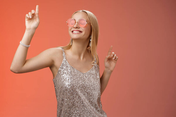 Восхитительный беззаботный привлекательный стильный миллениальный блондинка празднует веселье носить солнцезащитные очки модное серебряное платье танцы закрытые глаза широкая улыбка размахивая руками вверх, красный фон