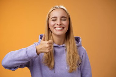 Neşeli, iyimser genç, heyecanlı, kapüşonlu, sarışın, kapüşonlu kadın model, geniş, kapalı gözlerle gülümseyen, eğlenceli, güzel jestler yapan harika tatil fikri, turuncu arka plan.