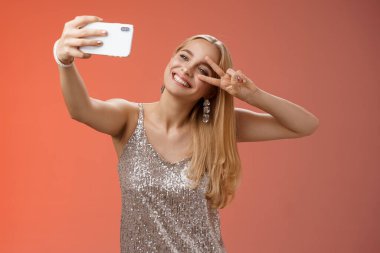 Şık, alımlı, pırıl pırıl, gümüş elbiseli, başı öne eğilmiş genç sarışın kadın barış jesti işareti kol kola uzanıp akıllı telefon tutarak selfie video kaydını çevrimiçi yapıyor.