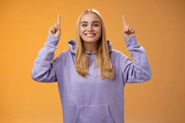 Дружня харизматична енергійна молода блондинка жінка в затишному худі піднімає руки, показуючи вказівні пальці, представляючи приголомшливу рекламу, яка посміхається з радістю рекомендувати продукт, помаранчевий фон — стокове фото