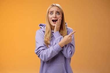 Şaşırtıcı derecede heyecanlı kapüşonlu genç sarışın kız avucunun içi gibi bağırıyor sağa doğru hayret verici bir şekilde haykırıyor ünlü süper star neşeli, pozitif duyguları tutamıyor, turuncu arka plan