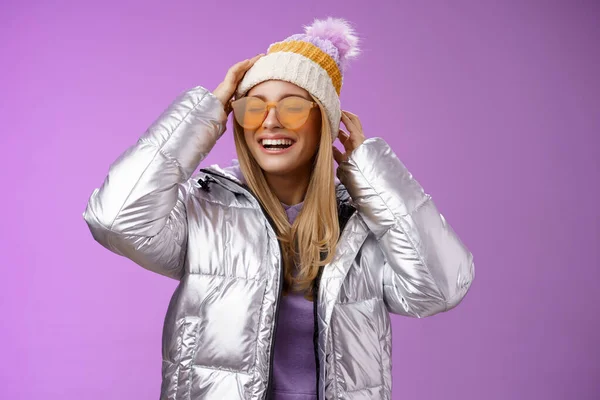 Ξανθιά κοπέλα που διασκεδάζει απολαμβάνοντας υπέροχες ηλιόλουστες χειμερινές διακοπές στο χιονοδρομικό κέντρο φορώντας γυαλιά ηλίου ασημί κομψό σακάκι με καπέλο χαμογελώντας χαρούμενα, μωβ φόντο — Φωτογραφία Αρχείου