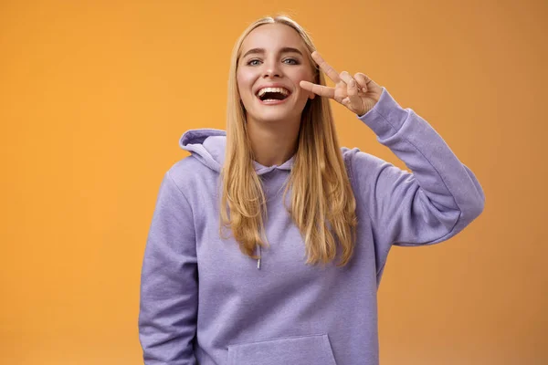 Ελκυστική φιλική ανέμελη χιλιετή ξανθιά κοπέλα με μωβ κουκούλα διασκεδάζοντας φίλοι γελώντας χαρούμενα δείχνουν ειρήνη νίκη hippie σημάδι λατρεύουν τέλεια ψυχρό καιρό άνοιξη, πορτοκαλί φόντο — Φωτογραφία Αρχείου