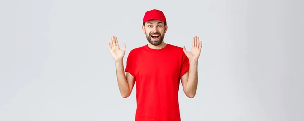 Online-Einkauf, Lieferung während der Quarantäne und Konzept zum Mitnehmen. Glücklich fröhlicher Kurier in rotem T-Shirt und Mütze, Firmenuniform, Hände hoch überrascht und amüsiert, stehend grauer Hintergrund — Stockfoto