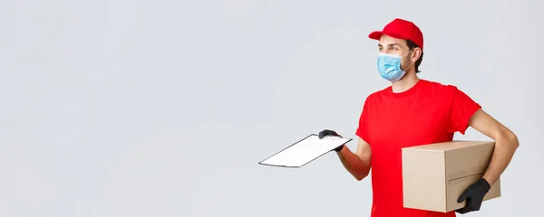 Packages and parcels delivery, covid-19 delivery, transfer orders. Profile of friendly courier in red uniform in face mask and gloves, bring order, hold box and give form for client to sign — Stock Photo, Image