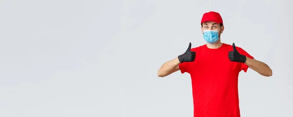 Covid-19, αυτο-καραντίνα, ψώνια και ναυτιλία έννοια. Ενθουσιαστικός και χαρούμενος κούριερ με κόκκινη στολή, γάντια και μάσκα προσώπου, ιδέα υποστήριξης, αντίχειρας-up, συνιστά ανέπαφη παράδοση πακέτου πελάτη — Φωτογραφία Αρχείου