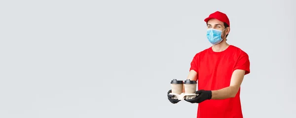 Takeaway, παράδοση τροφίμων και ειδών παντοπωλείου, covid-19 έννοια ανέπαφων παραγγελιών. Ευχάριστη κούριερ με κόκκινη στολή, μάσκα προσώπου και γάντια, που δίνει καφέ στον πελάτη, στέκεται γκρι φόντο — Φωτογραφία Αρχείου