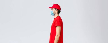 Covid-19, kendini karantina, online alışveriş ve nakliye konsepti. Posta şirketinin kırmızı üniformalı ciddi genç kuryesinin profili koruyucu tıbbi maske takmak, iş teslimatı, paket göndermek.