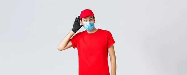 Groceries and packages delivery, covid-19, quarantine and shopping concept. Intrigued courier in red uniform, face mask and gloves, overhearing conversation, eavesdrop, hold hand near ear — Stock Photo, Image