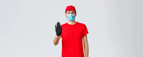 Covid-19, self-quarantine, online shopping concept. Confident delivery man in red uniform, gloves and face mask, raise hand in stop sign, prevent client step to storage without protective equipment — Stock Photo, Image