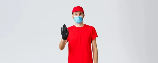 Covid-19, auto-cuarentena, concepto de compras en línea. Repartidor de confianza hombre en uniforme rojo, guantes y mascarilla, levante la mano en señal de stop, evitar que el cliente paso al almacenamiento sin equipo de protección —  Fotos de Stock