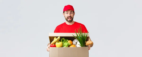 Groceries and packages delivery, covid-19, quarantine and shopping concept. Smiling handsome bearded courier in red uniform, bring food package, grocery order to client in box, look amused — Stock Photo, Image