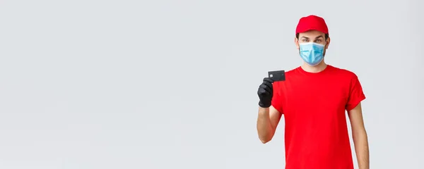 Contactless delivery, payment and online shopping during covid-19, self-quarantine. Young courier in red uniform cap, face mask and gloves, showing credit card, easy paying and ordering — Stock Photo, Image