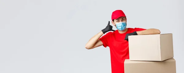 Packages and parcels delivery, covid-19 quarantine and transfer orders. Friendly courier in red uniform, face mask and gloves asking customer give call, show phone sign, stand near boxes — Stock Photo, Image