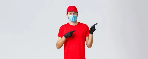 Covid-19, самокарантин, концепція інтернет-торгівлі та доставки. Заінтригований посміхнений хлопець доставки в червоній футболці, уніформа кеп-носія, використовуючи медичну маску і рукавички для доставки клієнту, вкажіть праворуч — стокове фото